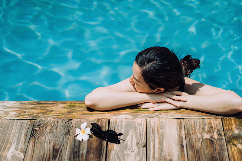 年轻的亚洲女人休息在游泳池的边缘放松和享受阳光，而在暑假图片素材