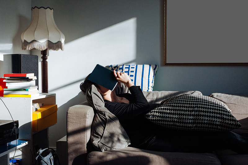 下午，一个年轻女子用一本书遮住脸睡在沙发上图片素材