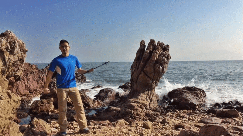 一名男子站在岩石海岸的拳头形状的岩石旁边，以汹涌的海浪为背景图片下载