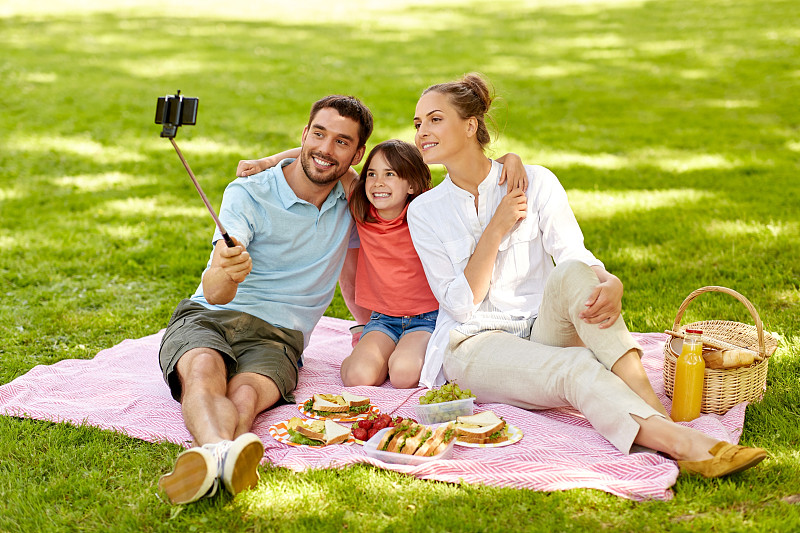 家庭，休闲和科技概念-快乐的母亲，父亲和女儿野餐和拍照的智能手机在夏季公园的自拍杆。一家人在公园野餐和自拍图片下载