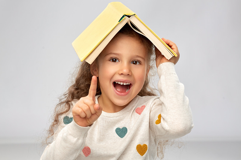 阅读，教育和童年的概念-肖像的微笑小女孩与书在屋顶上在灰色的背景。头戴书微笑女孩的肖像图片素材