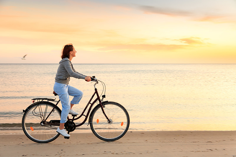 成熟的女人在日落的海滩上骑自行车图片素材