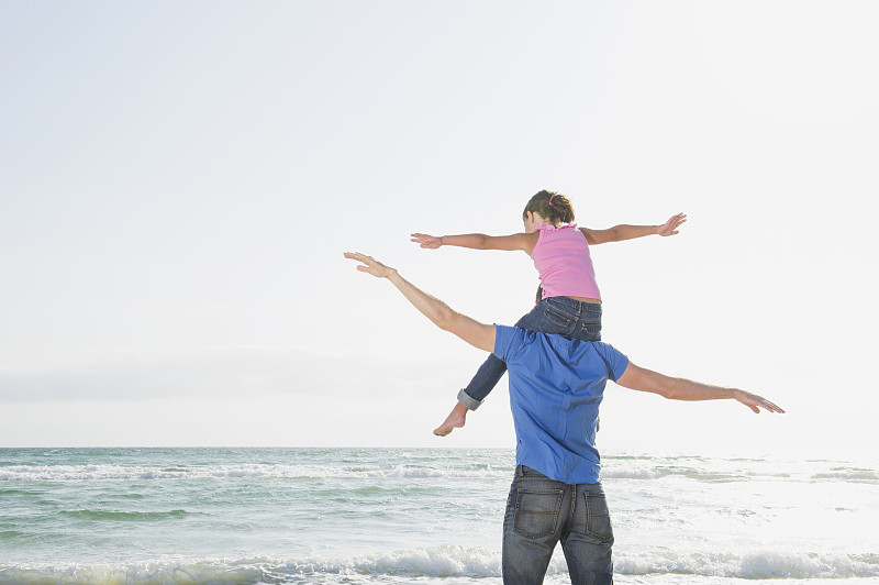 一个白人父亲肩上扛着一个会飞的女儿在海滩上图片下载