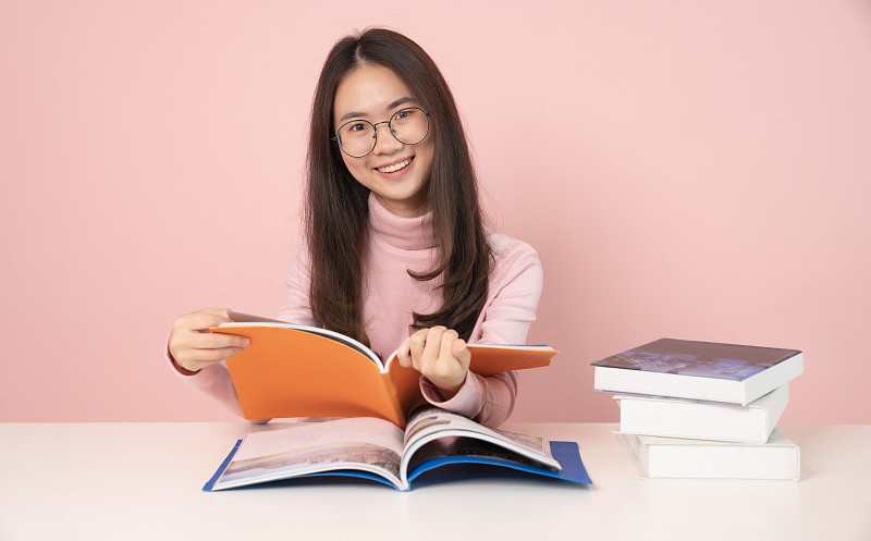 学习与教育理念。回归学校教育知识学院大学理念。美丽的女大学生拿着她的书，愉快地笑着坐在书桌上粉红色的背景图片素材