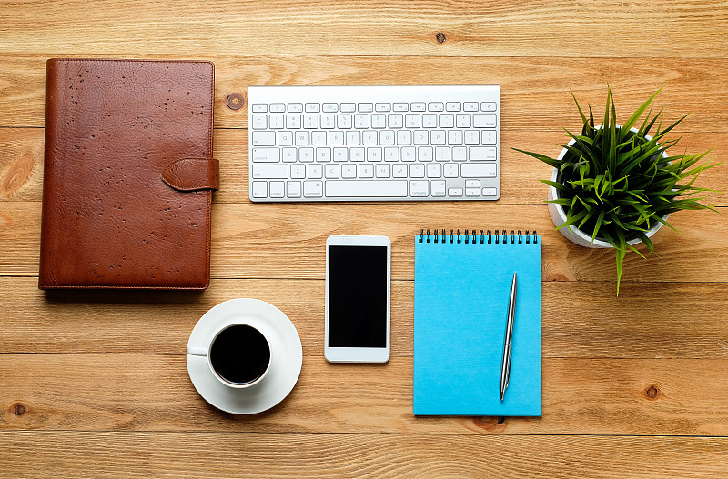 一个手机，一个电脑键盘，一支笔和记事本，一个咖啡杯和一朵花在木桌上。一个商人或经理在工作场所的工作主题。图片素材