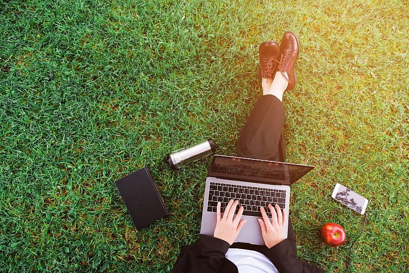 公园里的一个女孩坐在草地上用笔记本电脑工作，一个女人的手放在键盘上。靠近手机，苹果，笔记本。学习在户外。远程学习的概念图片下载