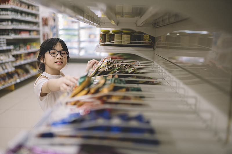 一个亚洲的中国小女孩从货架上挑选商品，然后在超市里拿起其中的一个图片下载