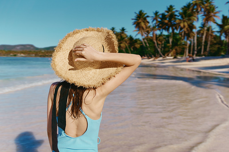 在一个热带岛屿上，一名身穿泳衣的年轻女子手戴一顶草帽，沿着沙滩散步，背景是棕榈树图片素材