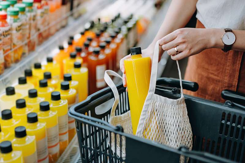 裁剪的镜头，年轻的亚洲妇女购物新鲜果汁冷藏货架在超市和把一瓶新鲜榨橙汁棉花网生态袋在购物车。“零浪费”的概念图片素材