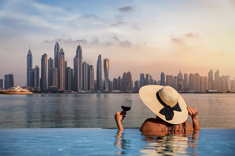 一位戴着帽子的妇女站在泳池边，欣赏着迪拜码头的天际线图片下载