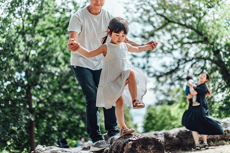 在一个阳光明媚的日子里，一位年轻的亚洲父亲牵着他的小女儿的手，扶着她沿着树干散步图片下载