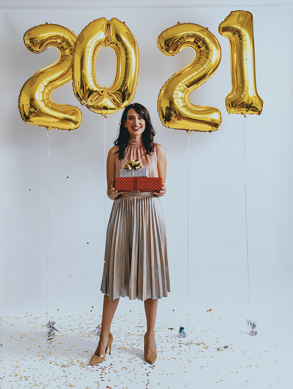 新的2021快乐!一个快乐的女人站在新年的气球下拿着礼物(看着相机)图片素材