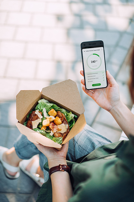 在肩的观点，年轻的亚洲商业女性使用智能手机上的健康和健身计划移动应用程序来定制她的日常饮食计划，检查营养事实和热量摄入的食物，而在城市公园吃沙拉午餐盒图片下载