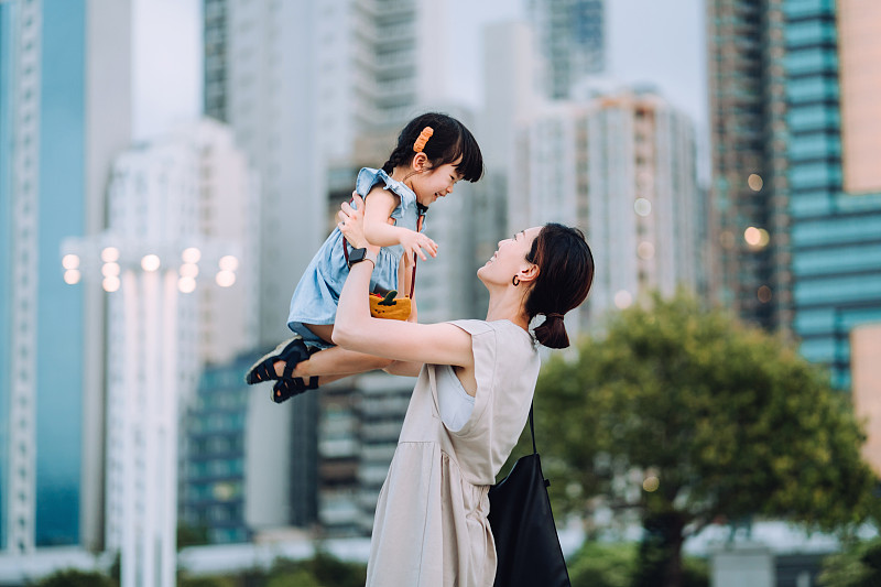 在公园里，一位可爱的年轻亚洲母亲将可爱的小女儿举到空中，映衬着城市的天际线。他们互相看着，笑得很开心。享受亲密的家庭时光图片下载