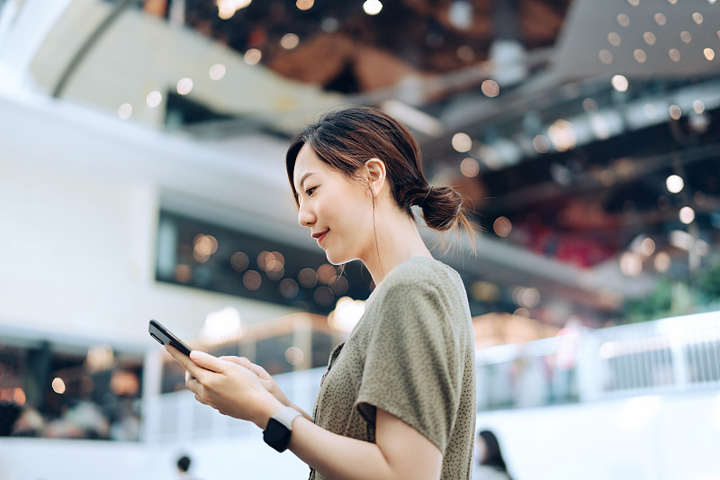 微笑的年轻亚洲女子在购物中心使用智能手机的侧面轮廓图片下载