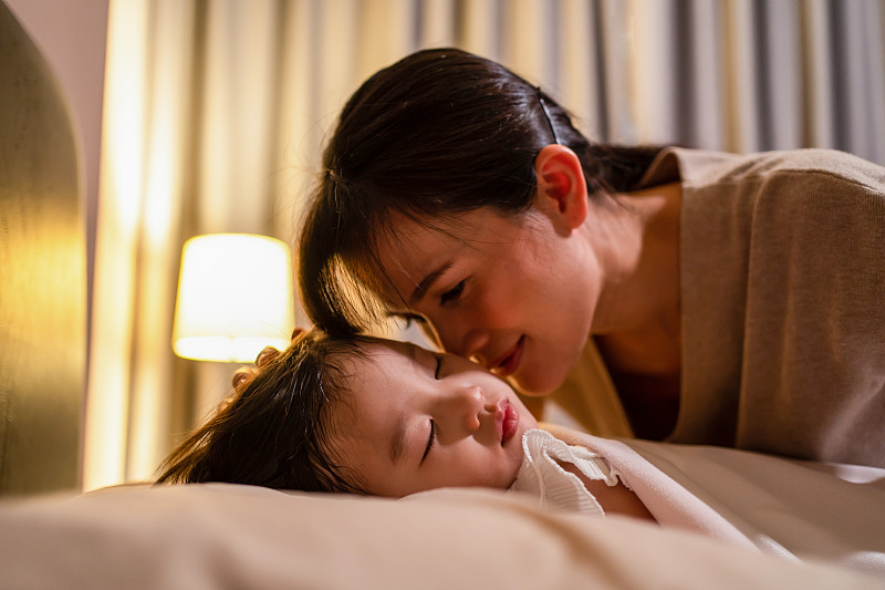 亚洲慈爱美丽的母亲温柔地吻在熟睡的幼小舒适的孩子的脸颊上。晚上，在家里黑暗的卧室里，慈母给熟睡的小女儿盖上毯子。图片下载
