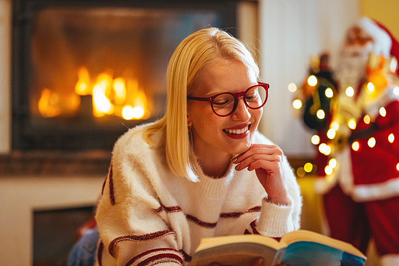 美丽的微笑金发女人拿着书，看着圣诞树。图片下载