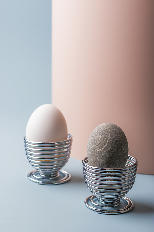 春节的概念是复活节。极简主义。新鲜的白色鸡蛋和石蛋在金属杯上的双粉红色和蓝色背景。垂直位置。本空间图片下载