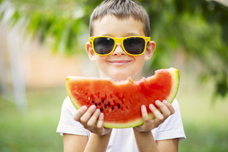 夏天戴着黄色墨镜的可爱男孩在户外吃西瓜。健康食用时令浆果和水果图片下载