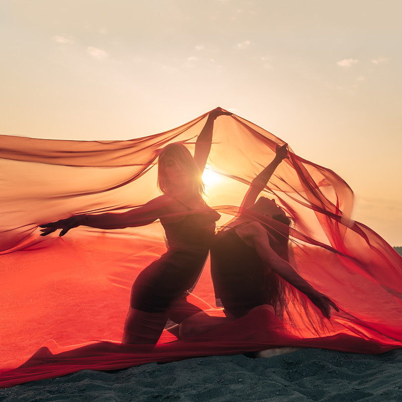 优雅的女孩在沙滩上表演舞蹈，在夕阳的映衬下，举起长长的红色织物。图片下载
