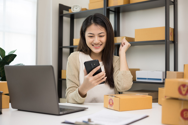 快乐年轻的亚洲女企业家在智能手机上检查产品。商务女性工作中小企业在线在家。请检查包裹上的地址。小企业主开始自由职业的概念。图片素材