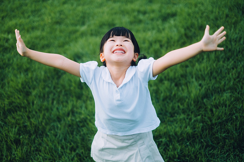 快乐的亚洲小女生在公园的草地上玩得很开心。她高兴地笑着，张开双臂。在夏日的户外享受大自然和自由时光。童年的生活方式。无忧无虑，自由和俏皮的概念图片下载