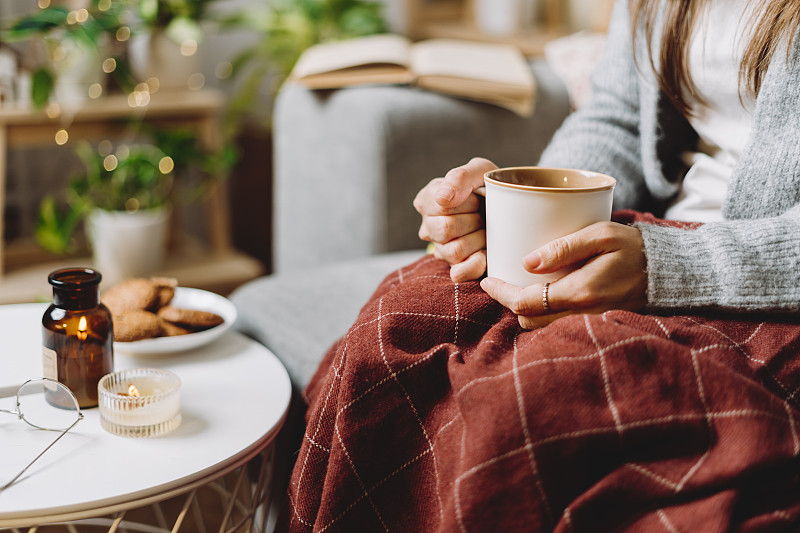舒适的女人腿针织冬天温暖的毛衣和格子格喝热可可或咖啡杯，坐在家里的沙发上。蜡烛、装饰、饼干和室内植物都是秋天的氛围。图片下载