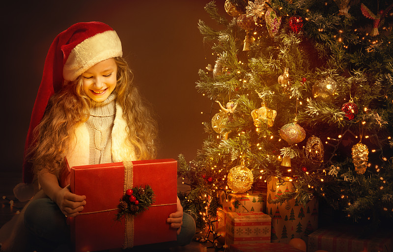 圣诞孩子打开礼物旁边的圣诞树。快乐的女孩在圣诞老人的帽子看里面的礼盒与闪耀的光在黑暗的家庭房间。圣诞夜魔法夜图片下载
