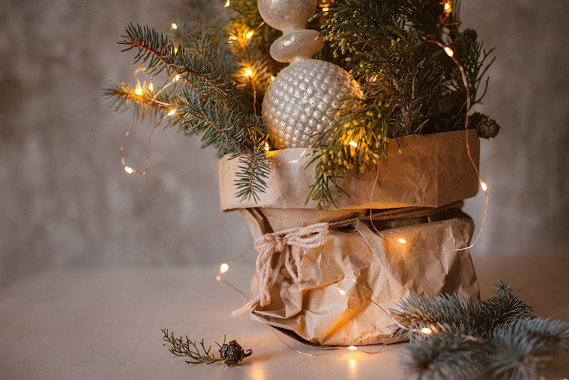 天然的圣诞花束装在工艺纸袋里，配上花环灯和玻璃顶顶装饰，衬着灰色的混凝土墙。图片下载