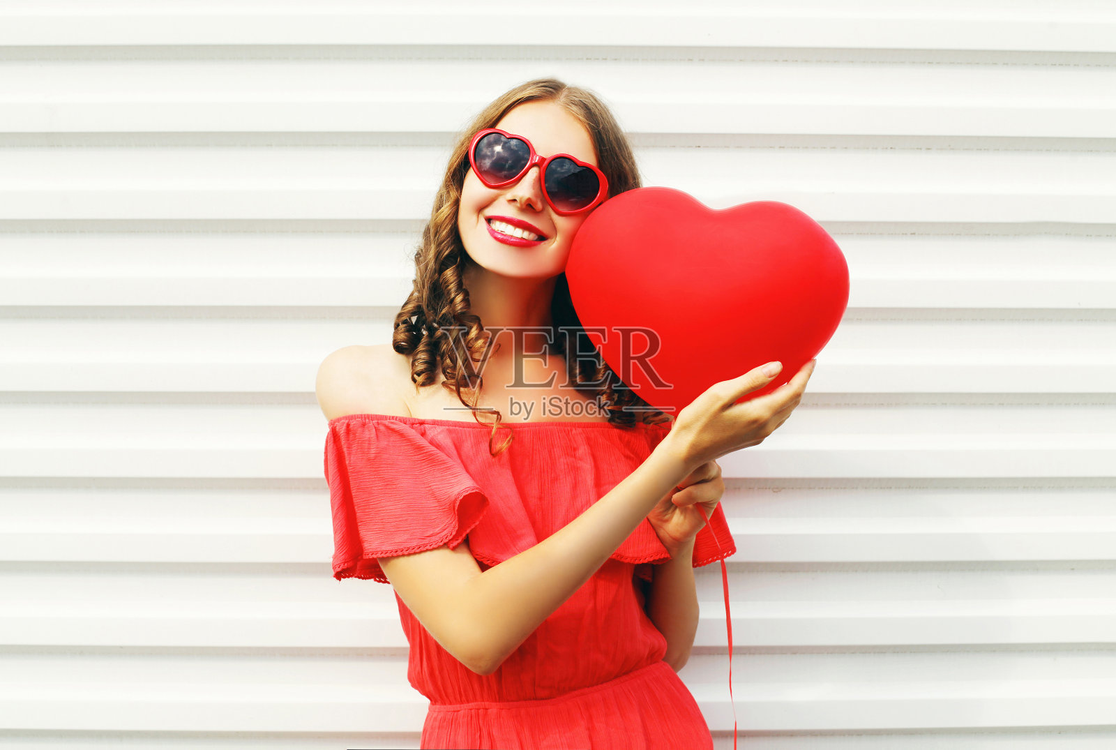 漂亮的微笑女人穿着红色的裙子和太阳眼镜与气球心脏形状的城市在白色的背景照片摄影图片