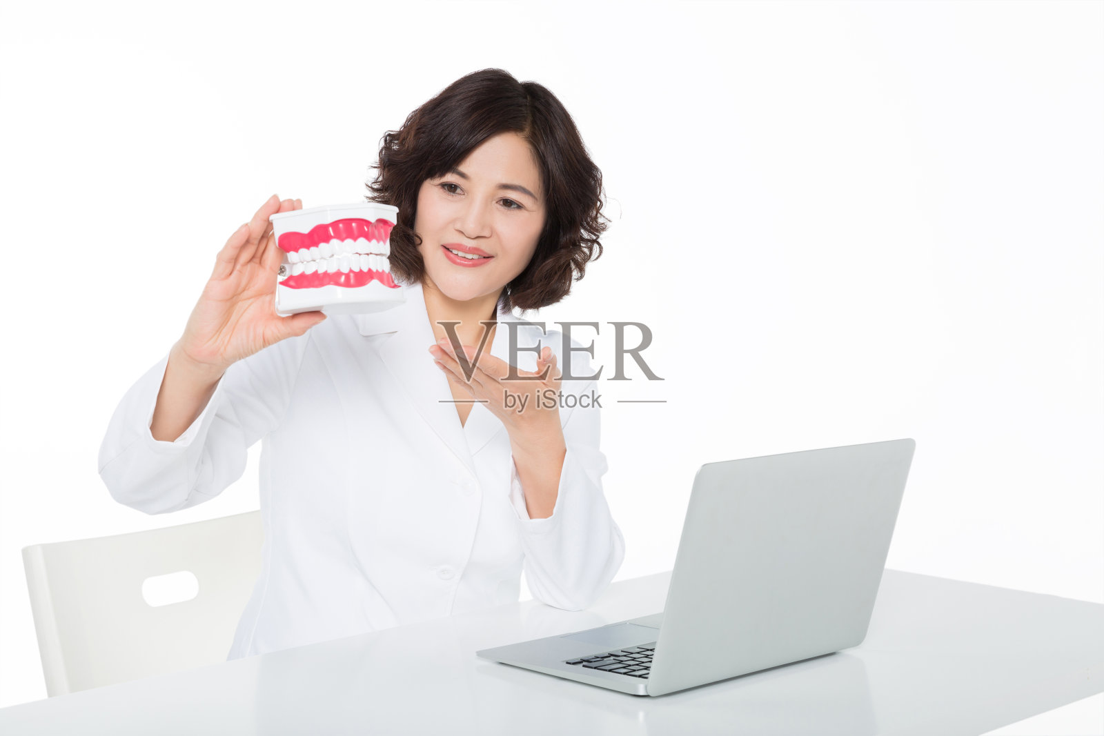 一个牙科医生拿着假牙，在白色背景的办公桌上放着一台笔记本电脑照片摄影图片