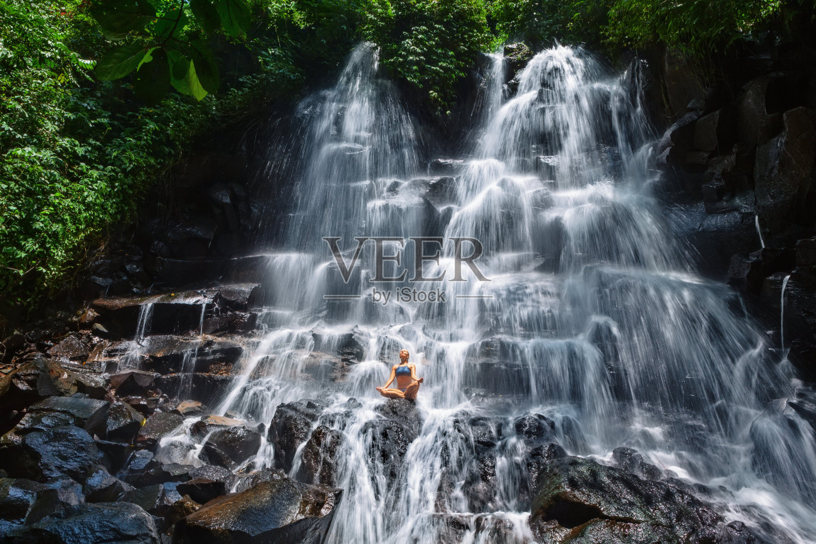 女人坐在岩石在瀑布下瑜伽姿势照片摄影图片