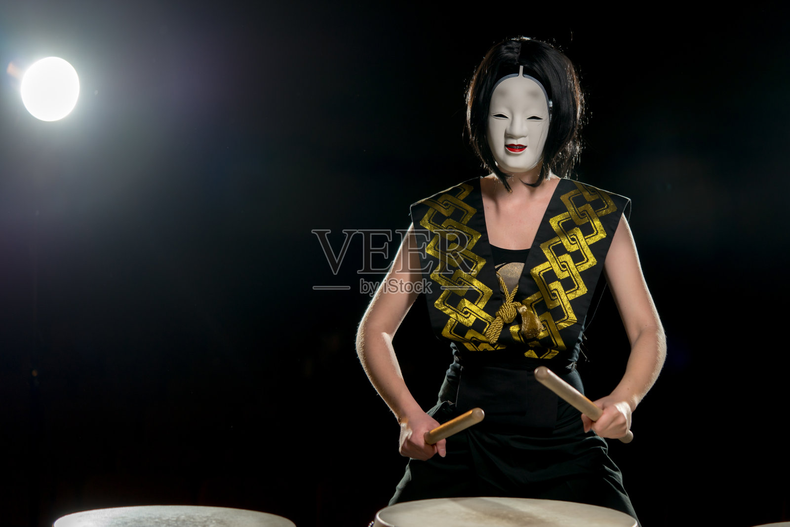 鼓手女孩在白色的恶魔面具和鼓槌，工作室拍摄在一个黑暗的背景。照片摄影图片