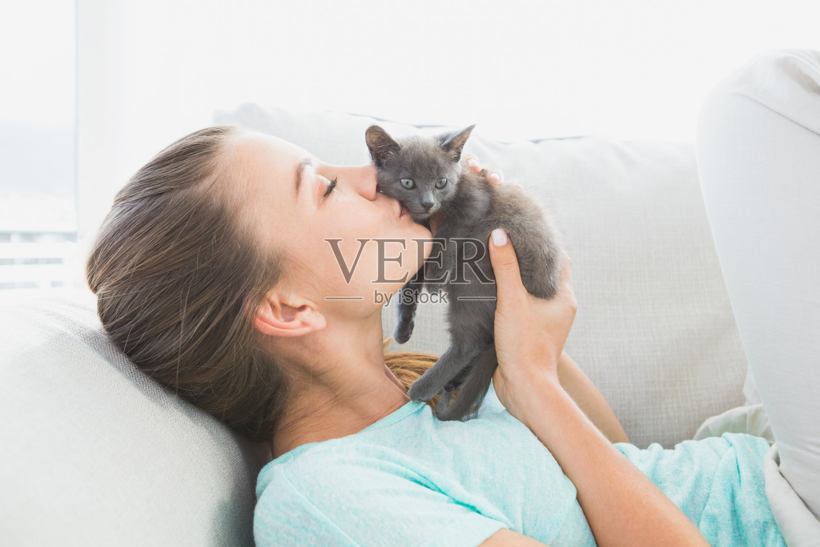 快乐的女人躺在沙发上亲吻一只灰色的小猫照片摄影图片