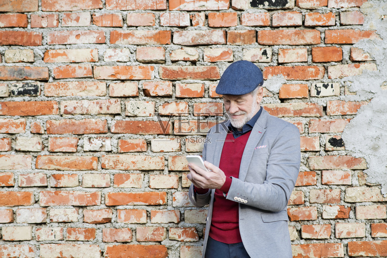 老男人拿着智能手机靠在砖墙上发短信。照片摄影图片