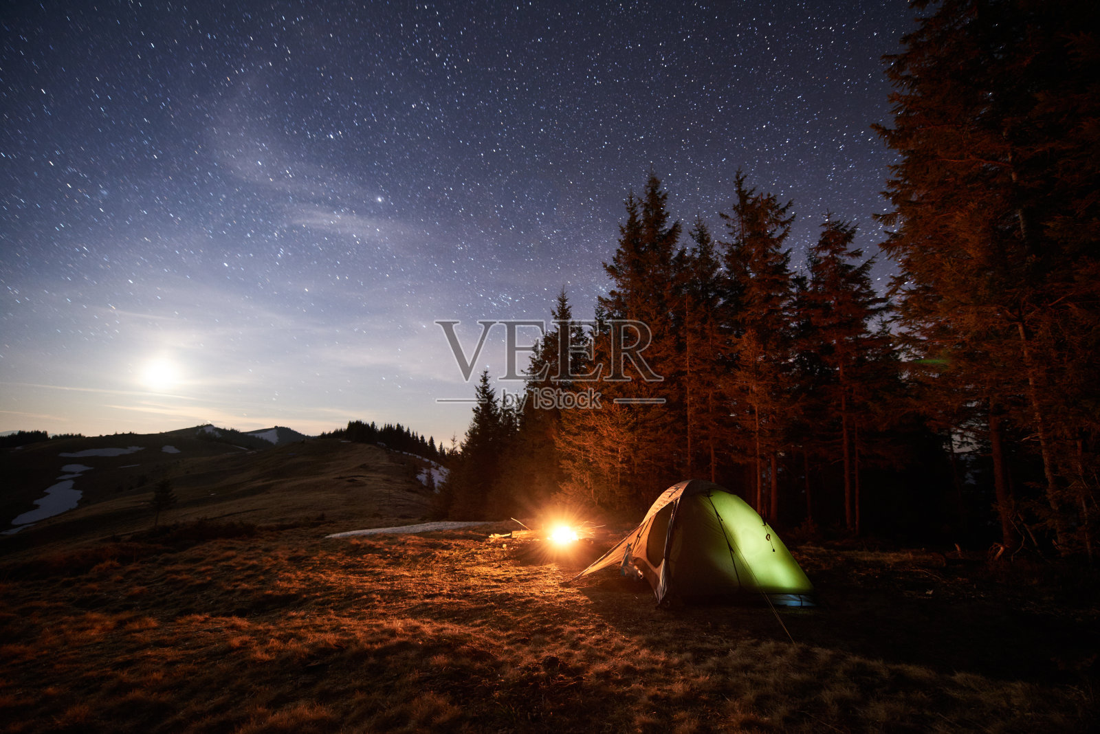 游客晚上在森林附近露营。在满是星星和月亮的美丽夜空下，照亮了帐篷和篝火照片摄影图片