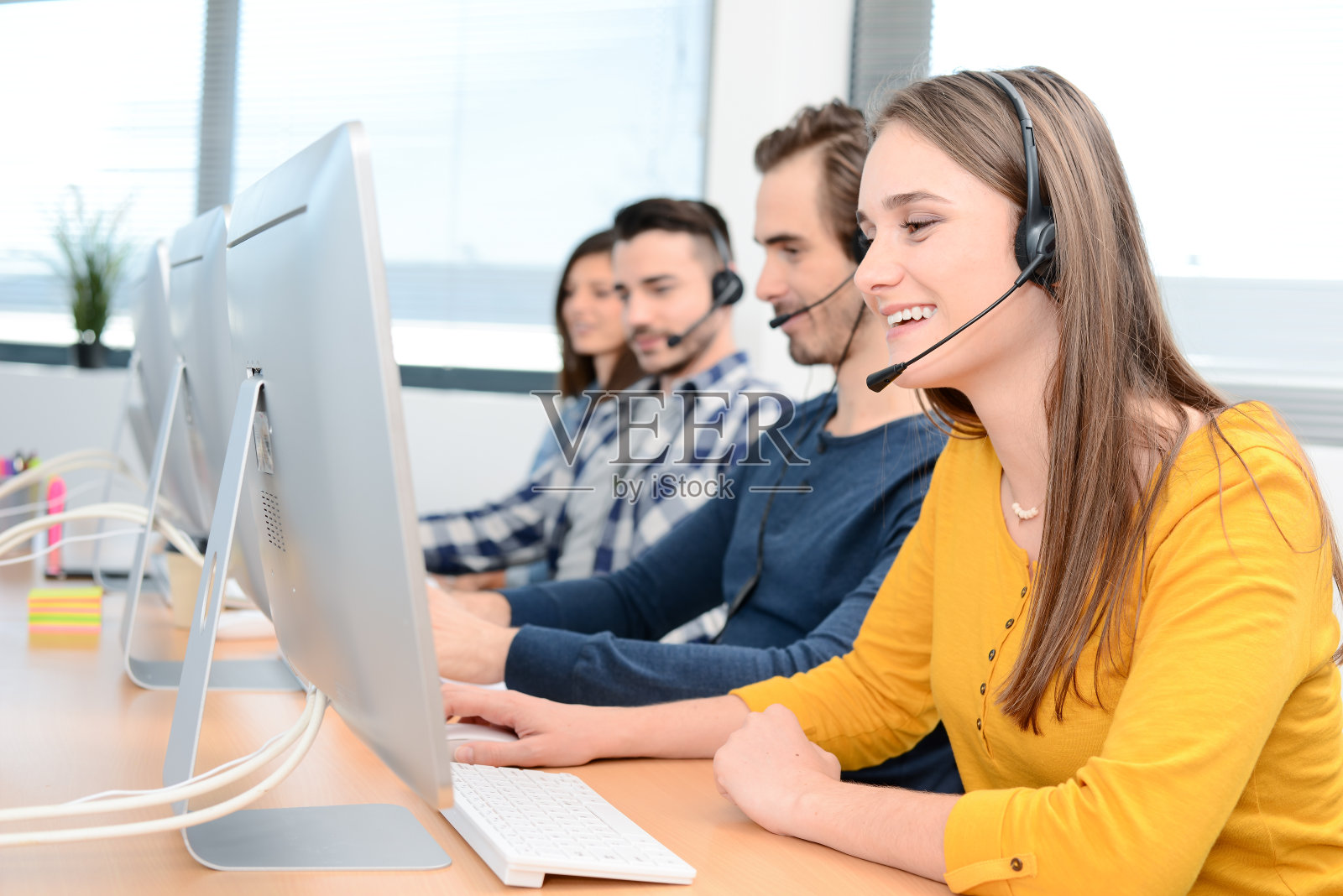 一个美丽开朗的年轻女性电话接线员戴着耳机在桌面电脑上工作，在客户服务呼叫支持热线业务中心，团队工作人员在后台照片摄影图片