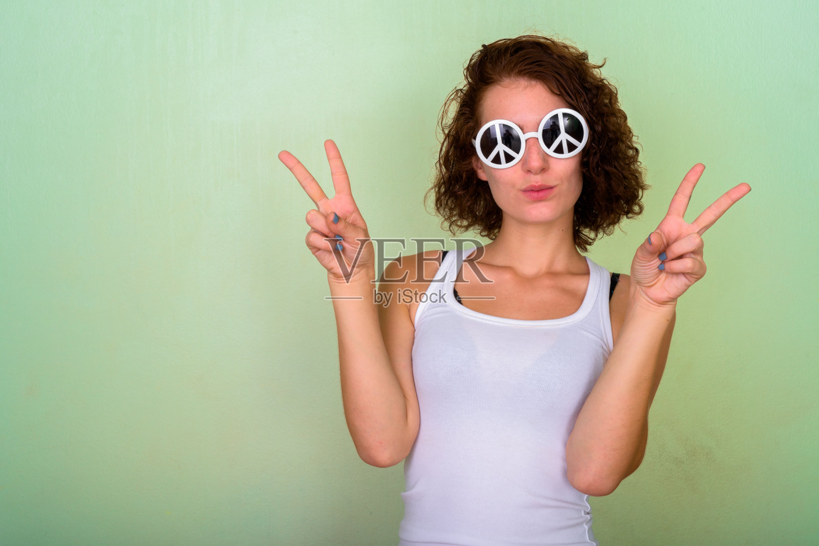 工作室拍摄的美丽的十几岁的女孩戴着太阳镜与和平的手势，同时给予和平的双手对着绿色的墙壁照片摄影图片