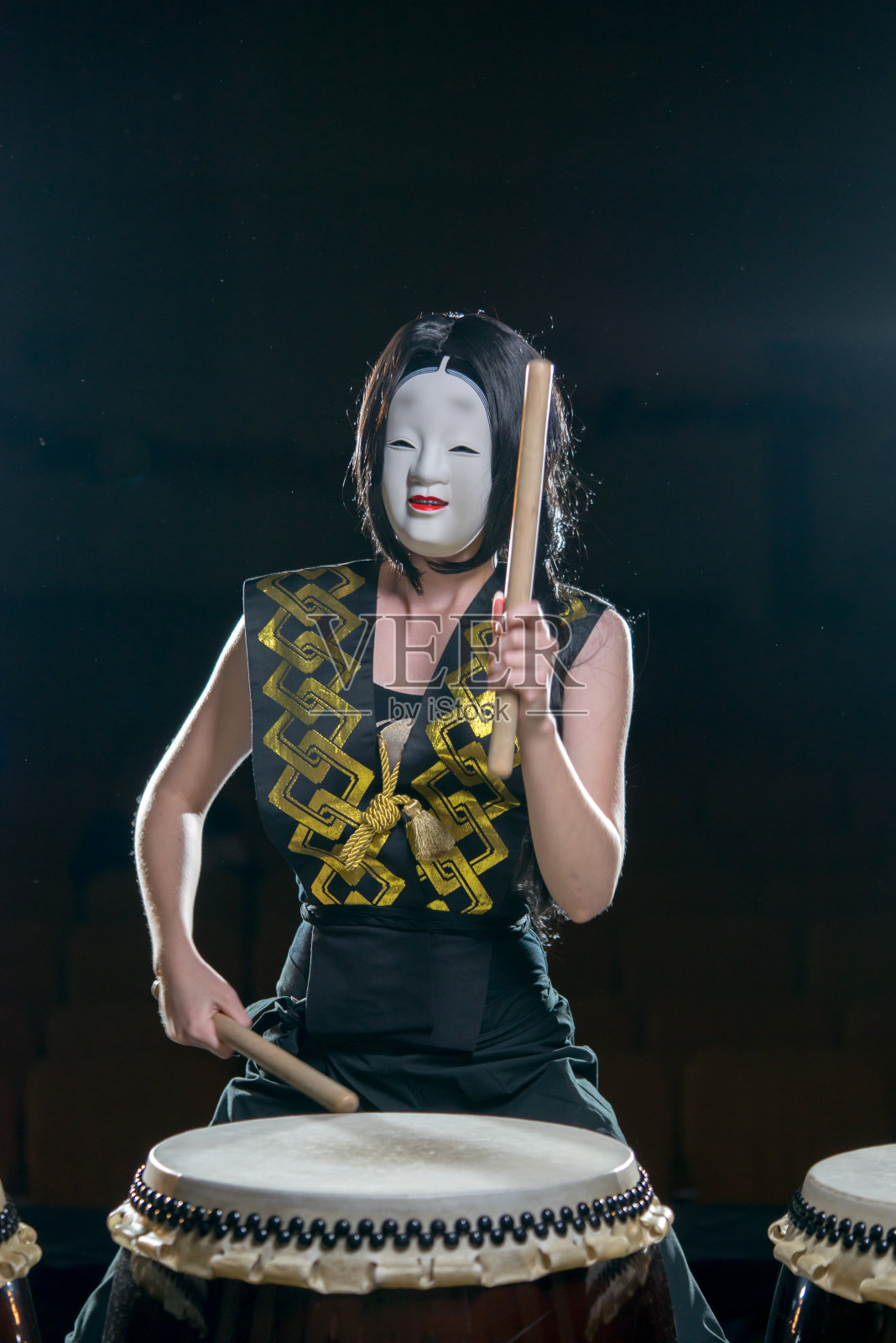 鼓手女孩在白色的恶魔面具和鼓槌，工作室拍摄在一个黑暗的背景。照片摄影图片
