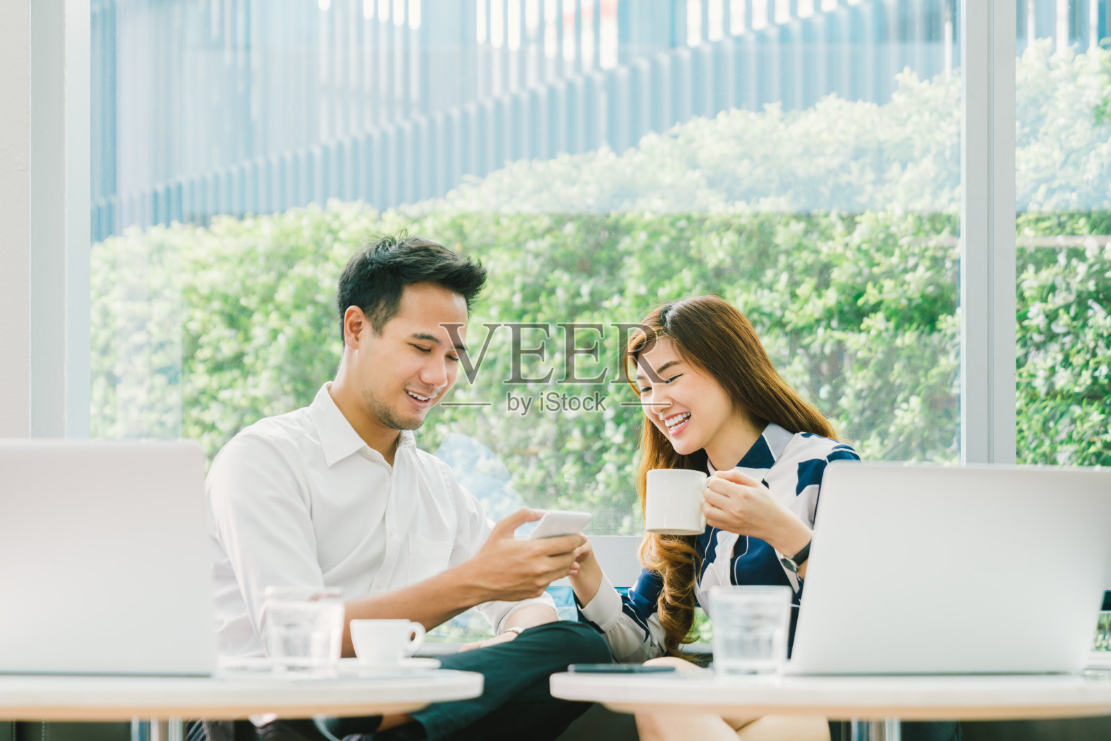 年轻的亚洲夫妇，同事，或商业伙伴有乐趣一起使用智能手机，与笔记本电脑在咖啡店。信息技术，咖啡生活方式，浪漫关系概念照片摄影图片