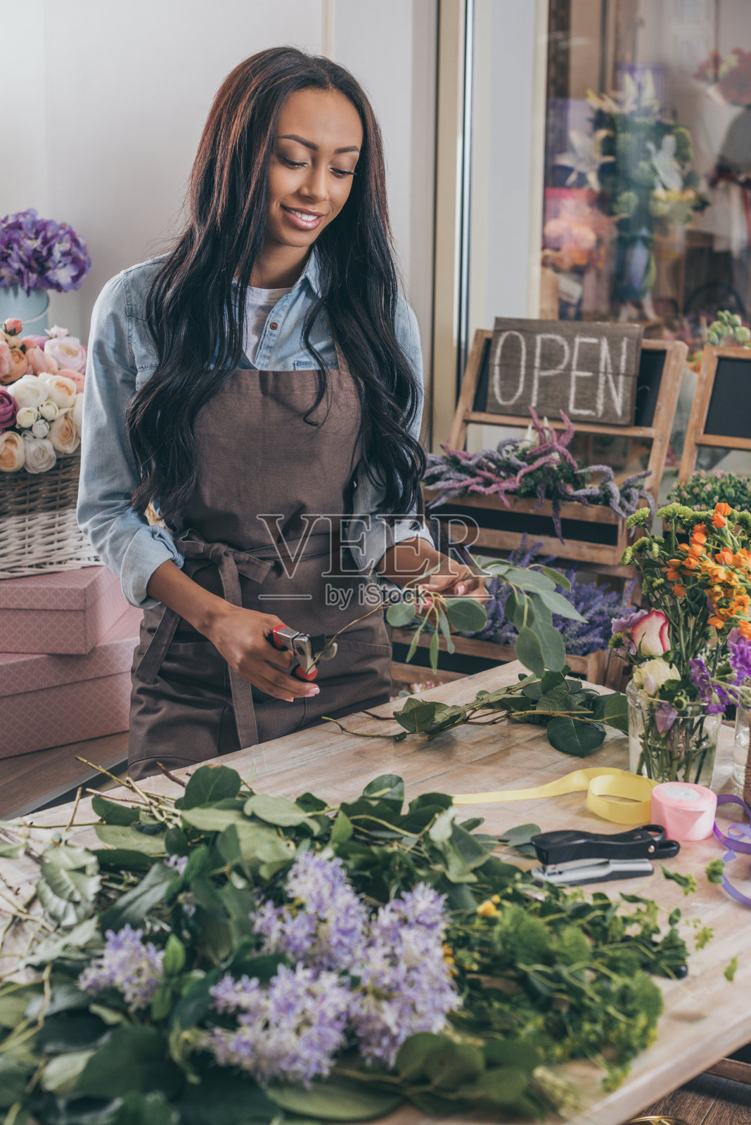 美丽微笑的非洲裔美国妇女在围裙拿着剪枝夹和工作与新鲜的植物在花店照片摄影图片
