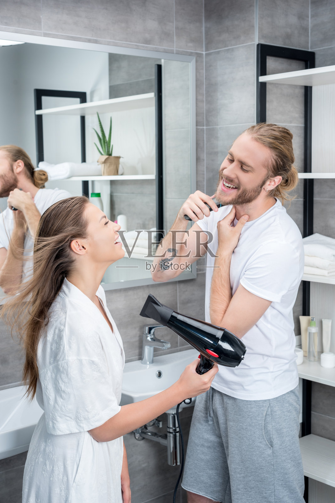 微笑的丈夫刮胡子，而妻子在浴室烘干头发在早上照片摄影图片