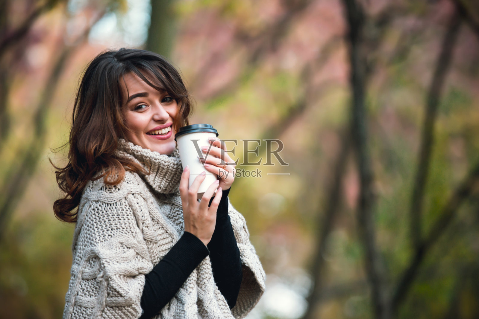 美丽的女孩穿着暖和的毛衣，在秋天的公园里喝着咖啡。温暖的阳光明媚的天气。在户外。照片摄影图片