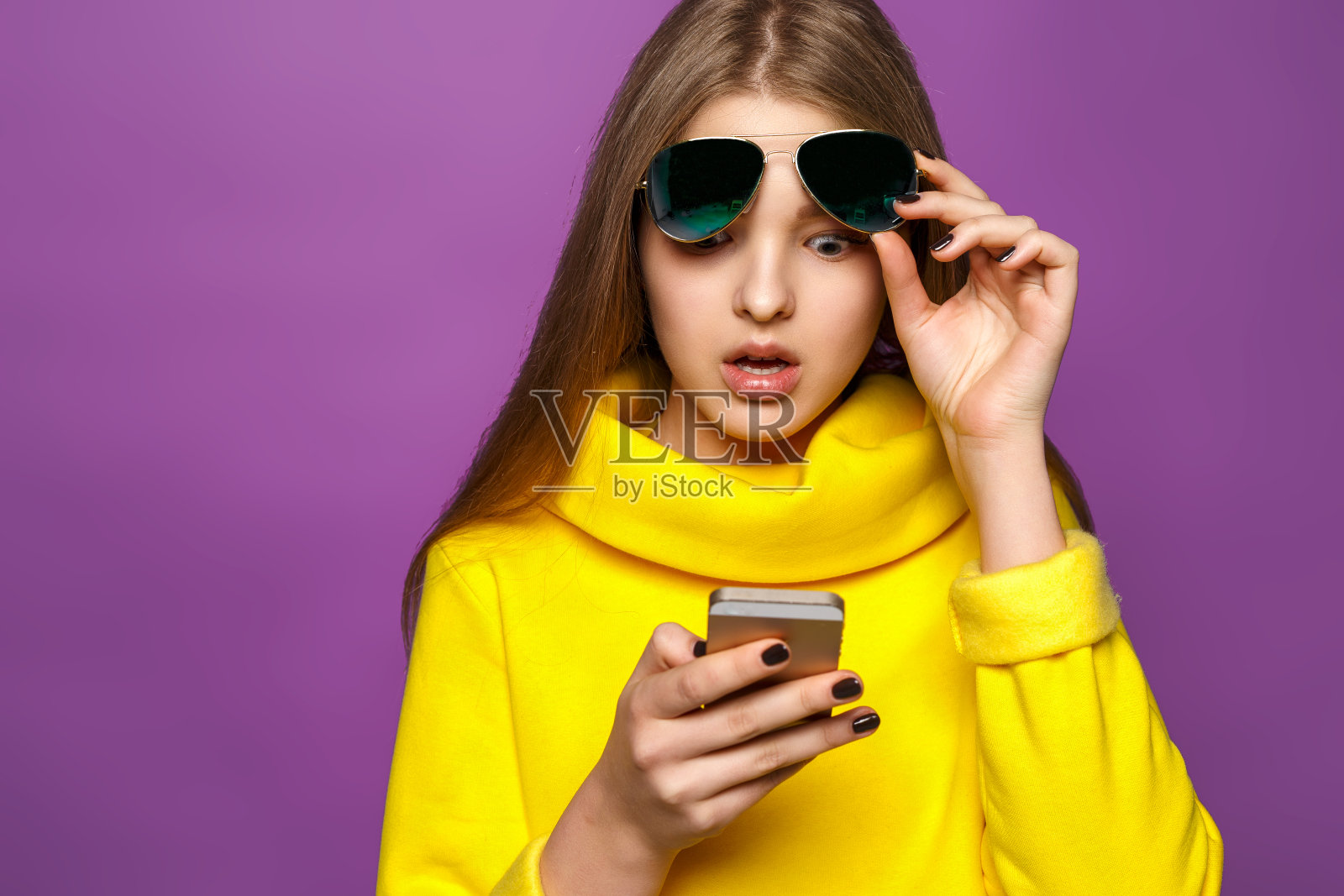 肖像惊讶的年轻女孩从智能手机短信在明亮的黄色毛衣，孤立在紫罗兰的背景照片摄影图片