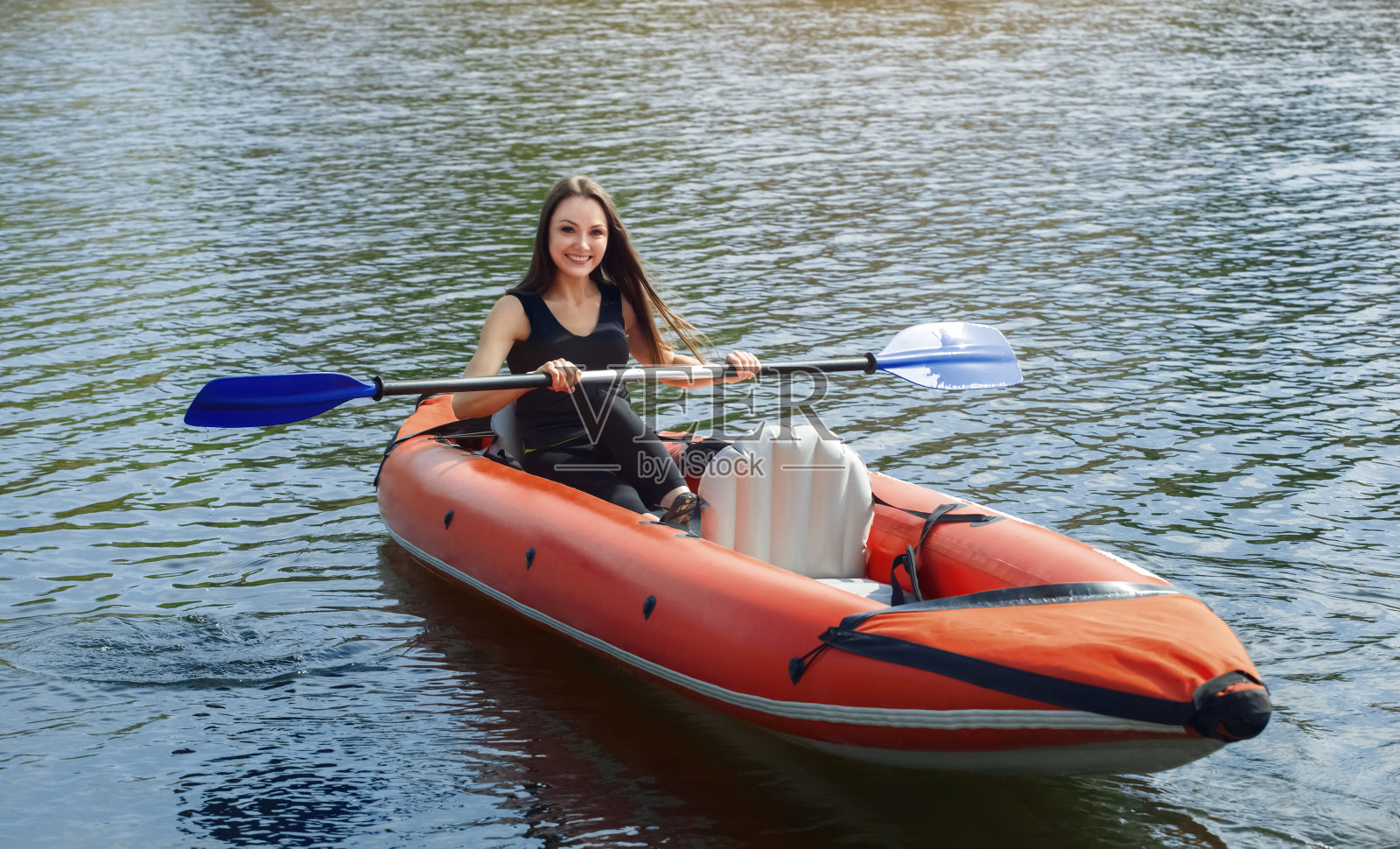 那个微笑着的女孩——一个长头发的女运动员，穿着黑色运动服，在一个温暖的夏日阳光明媚的日子里，在湖上的红色充气独木舟上划着桨照片摄影图片