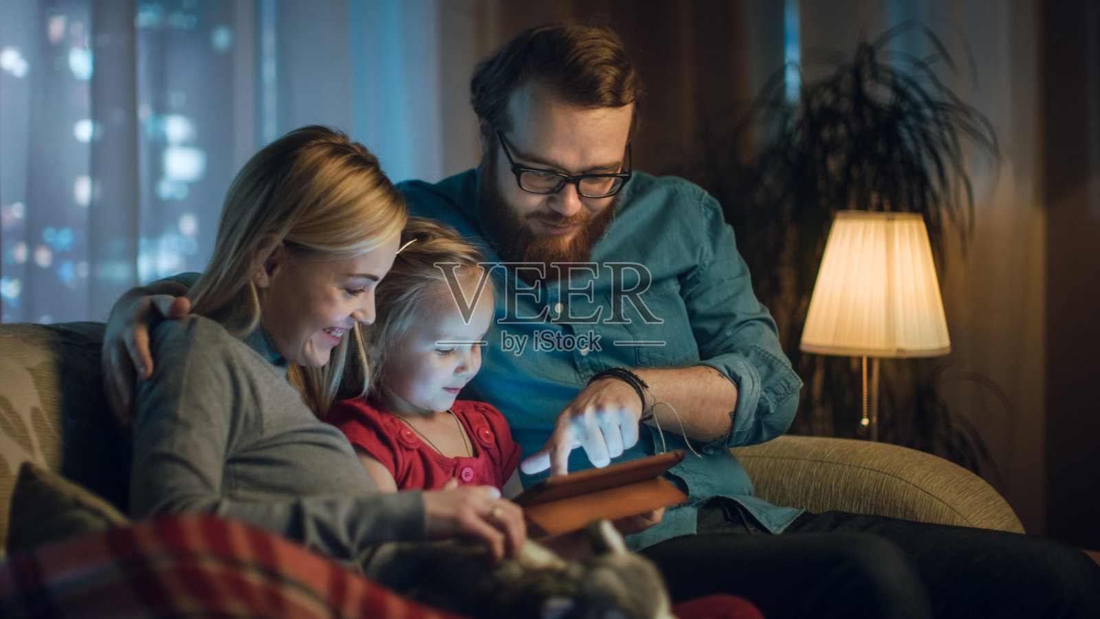爸爸、妈妈和小女儿在客厅的沙发上读儿童读物。这是晚上。照片摄影图片