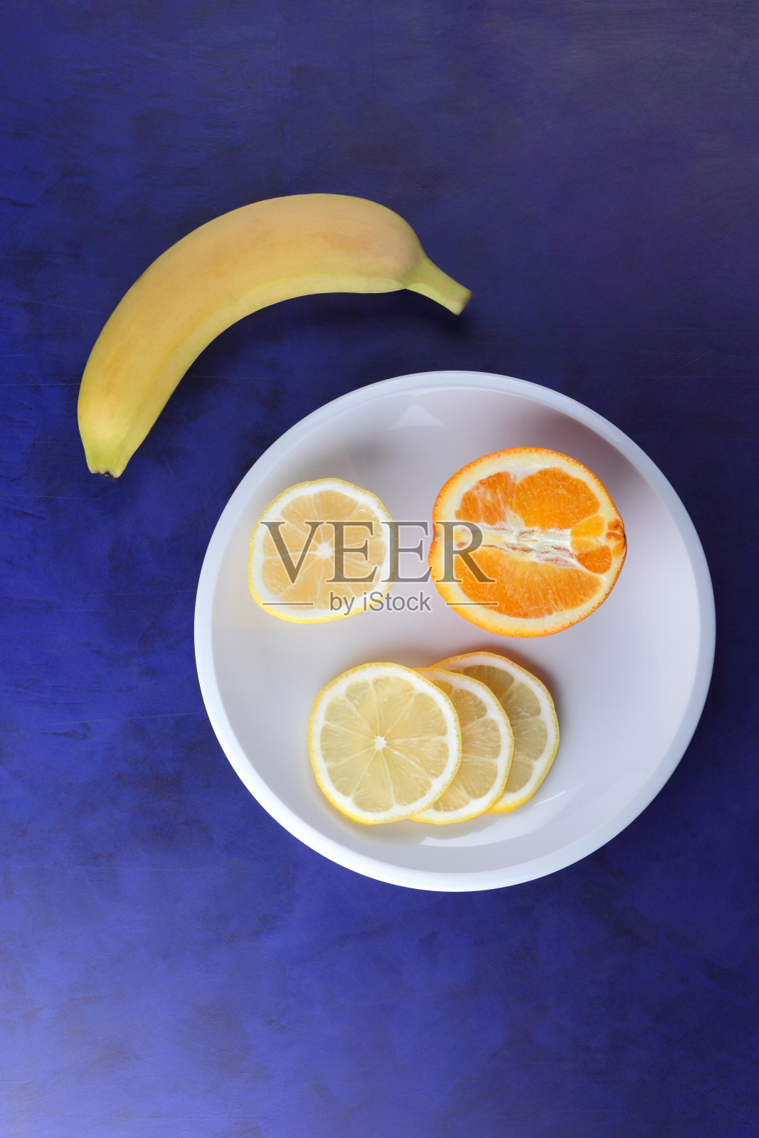 极简主义风格的紫外光背景柑橘，白色盘子上的柠檬片和半片橙子，深蓝色背景上的黄色香蕉，健康食品，素食，流行艺术照片摄影图片