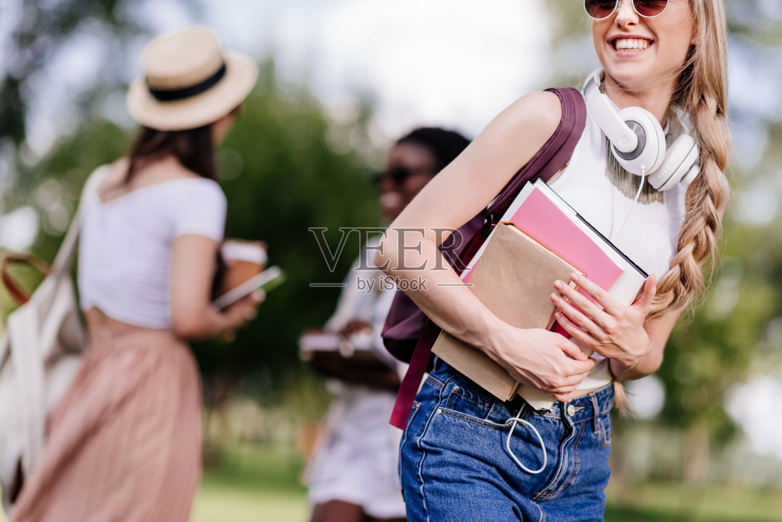 美丽微笑的金发学生戴着耳机拿着书，朋友们在公园后面聊天照片摄影图片