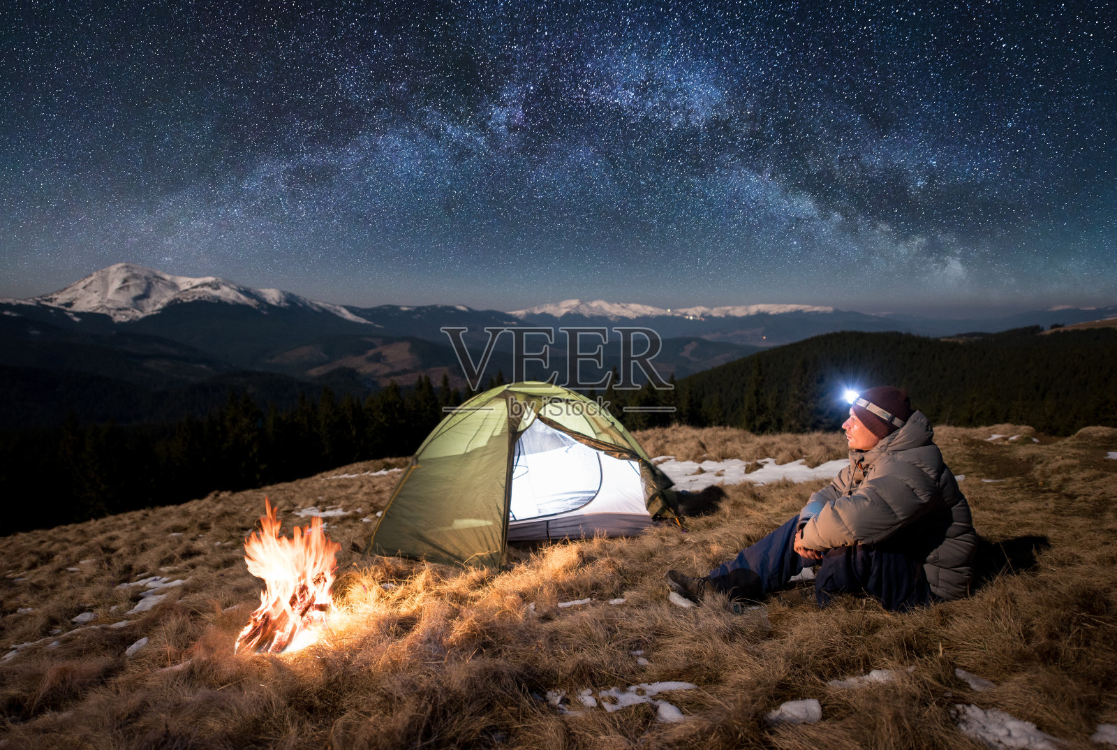 男游客晚上在营地休息。在满是星星和银河的美丽天空下，一个带着头灯的家伙坐在篝火和帐篷旁。背景是白雪皑皑的群山照片摄影图片