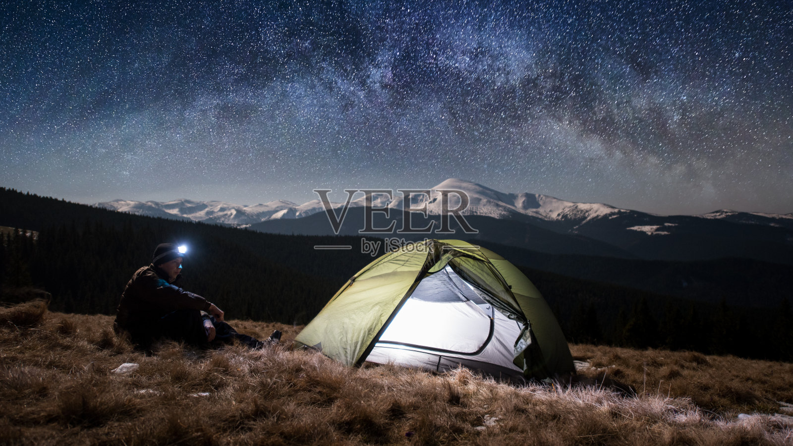 一名徒步旅行者晚上在山里露营，欣赏夜景。在满是星星和银河的美丽夜空下，一个带着头灯的男人坐在旅游帐篷附近。图片宽高比16:9照片摄影图片